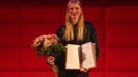 Eine Person mit langen blonden Haaren hält einen Blumenstrauß und eine aufgeschlagene Urkunde vor einem roten, gepolsterten Hintergrund.