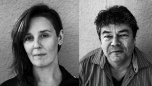 Zwei Personen, eine Frau und ein Mann mittleren Alters, blicken ernst in die Kamera vor neutralem Hintergrund.