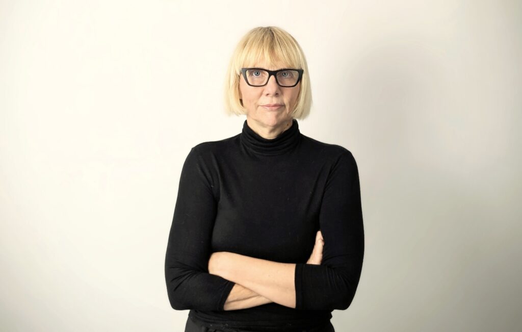 Frau mit blonden Haaren, Brille und schwarzem Rollkragenpullover steht vor hellem Hintergrund mit verschränkten Armen.