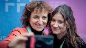 Zwei Frauen lächeln in die Kamera und machen ein Selfie vor einem blau-lila Hintergrund mit weißen Buchstaben.