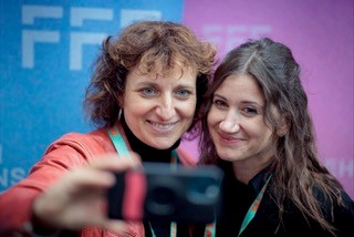 Zwei Frauen lächeln in die Kamera und machen ein Selfie vor einem blau-lila Hintergrund mit weißen Buchstaben.