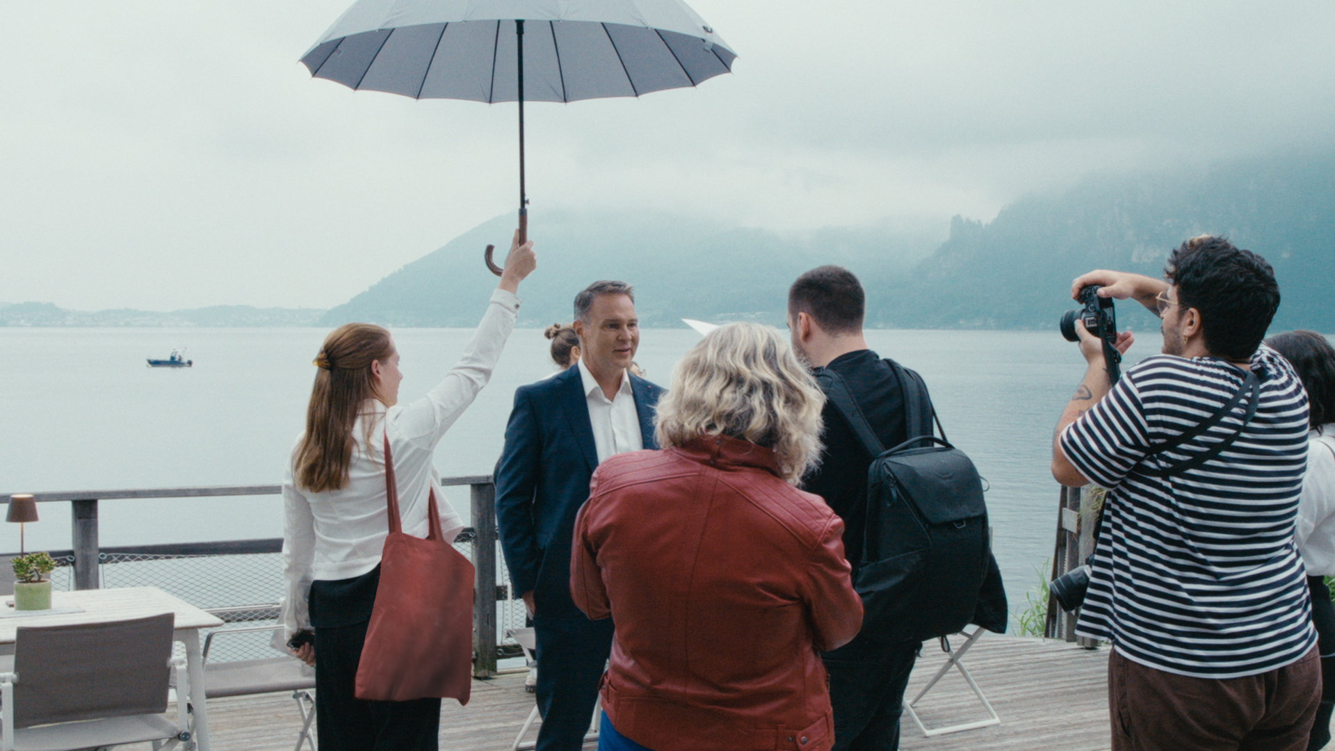 Eine Frau hält einem Mann im Anzug, Andreas Babler, einen Schirm, während mehrere Personen ihn am See mit Kameras fotografieren.
