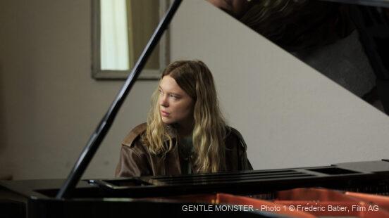Eine junge Frau mit langen blonden Haaren Lucy (gespielt von Léa Seydoux) sitzt nachdenklich an einem schwarzen Flügel in einem hellen Raum.