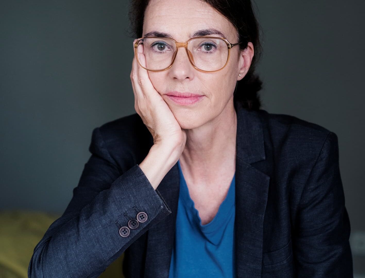 Frau mit Brille und dunklem Blazer sitzt nachdenklich mit Kopf auf der Hand gestützt vor neutralem Hintergrund.