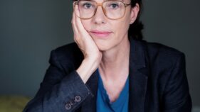 Frau mit Brille und dunklem Blazer sitzt nachdenklich mit Kopf auf der Hand gestützt vor neutralem Hintergrund.