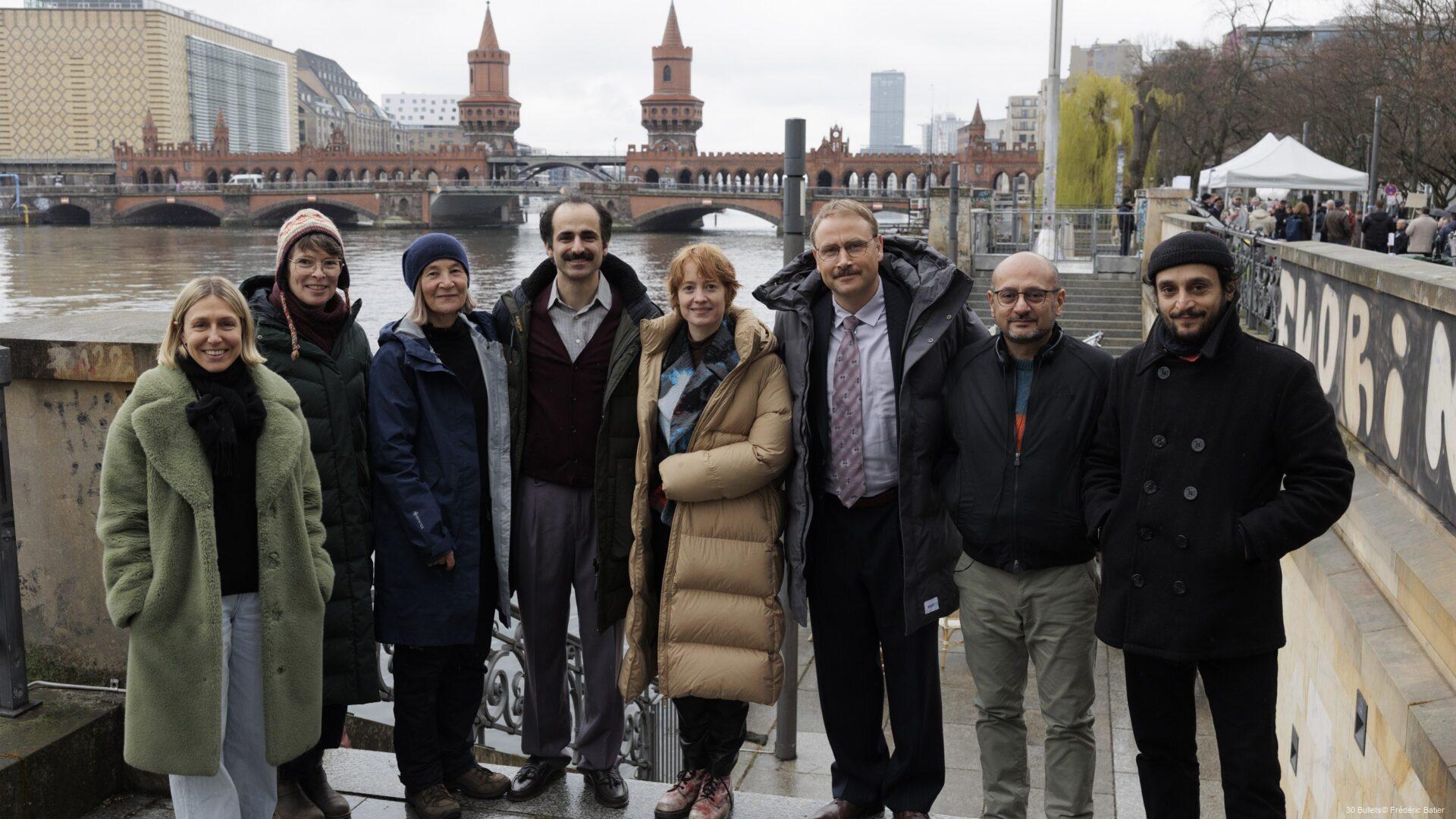Eine Gruppe von acht Menschen steht lachend an der Spree mit Blick auf die Oberbaumbrücke in Berlin bei bewölktem Wetter. Von links nach rechts: Judith Fülle (Produzentin), Solveig Cornelisen (ZDF), Judith Kaufmann (Kamera), Hadi Khanjanpour, Leonie Benesch, Max Riemelt, Arash T. Riahi (Drehbuch, Regie, Co-Produzent), Arman T. Riahi (Drehbuch, Regie)