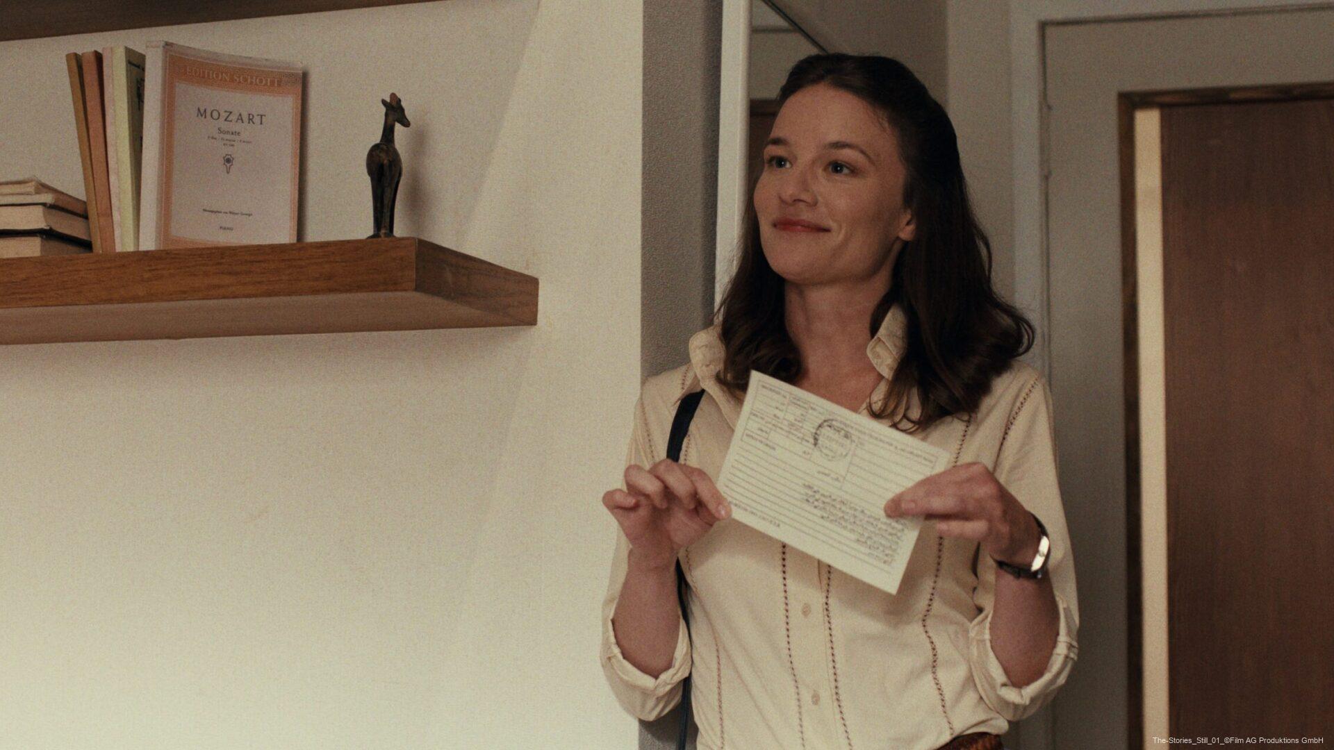 Frau steht lächelnd in einem Raum und hält ein Papier hoch; im Hintergrund Regal mit Büchern und einer Pferdeskulptur.