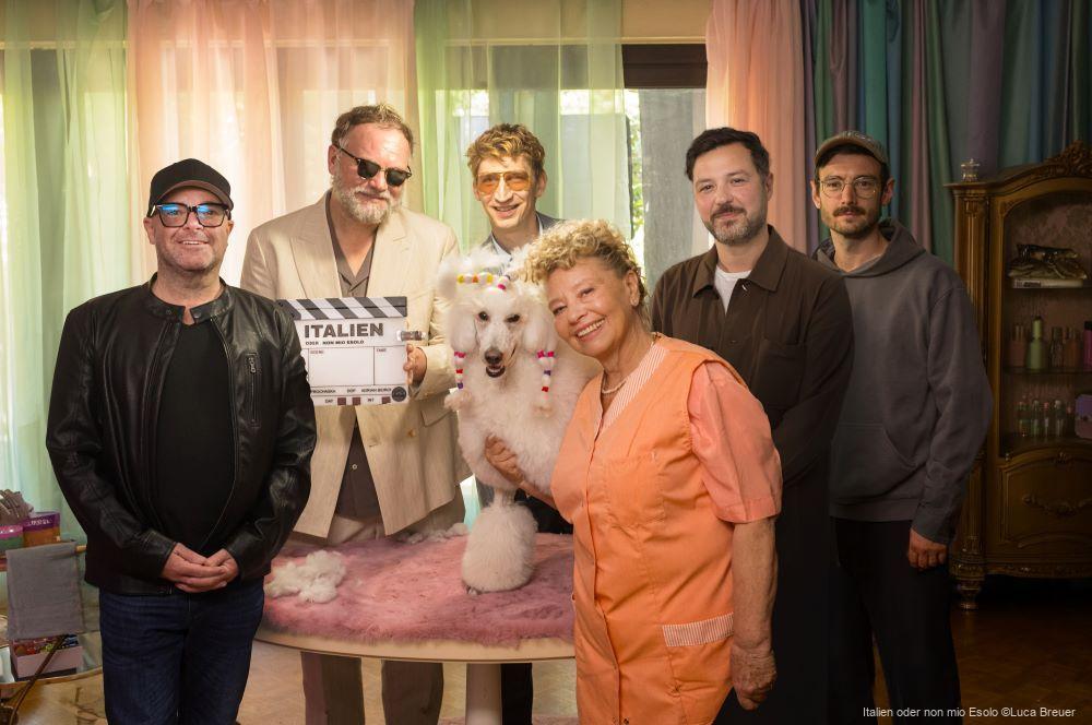 Eine Gruppe von sechs Erwachsenen und einem weißen Pudel steht lächelnd um einen Filmbrett mit der Aufschrift „Italien“ in einem bunten Raum. v.l.n.r: Markus Pauser, Thomas Stipsits, Lukas Walcher, Margarethe Tiesel, Daniel Prochaska, Adrian Bidron