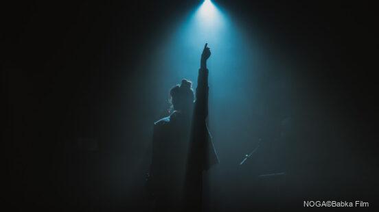 Silhouette einer Person auf einer dunklen Bühne, die im blauen Scheinwerferlicht den Arm nach oben streckt