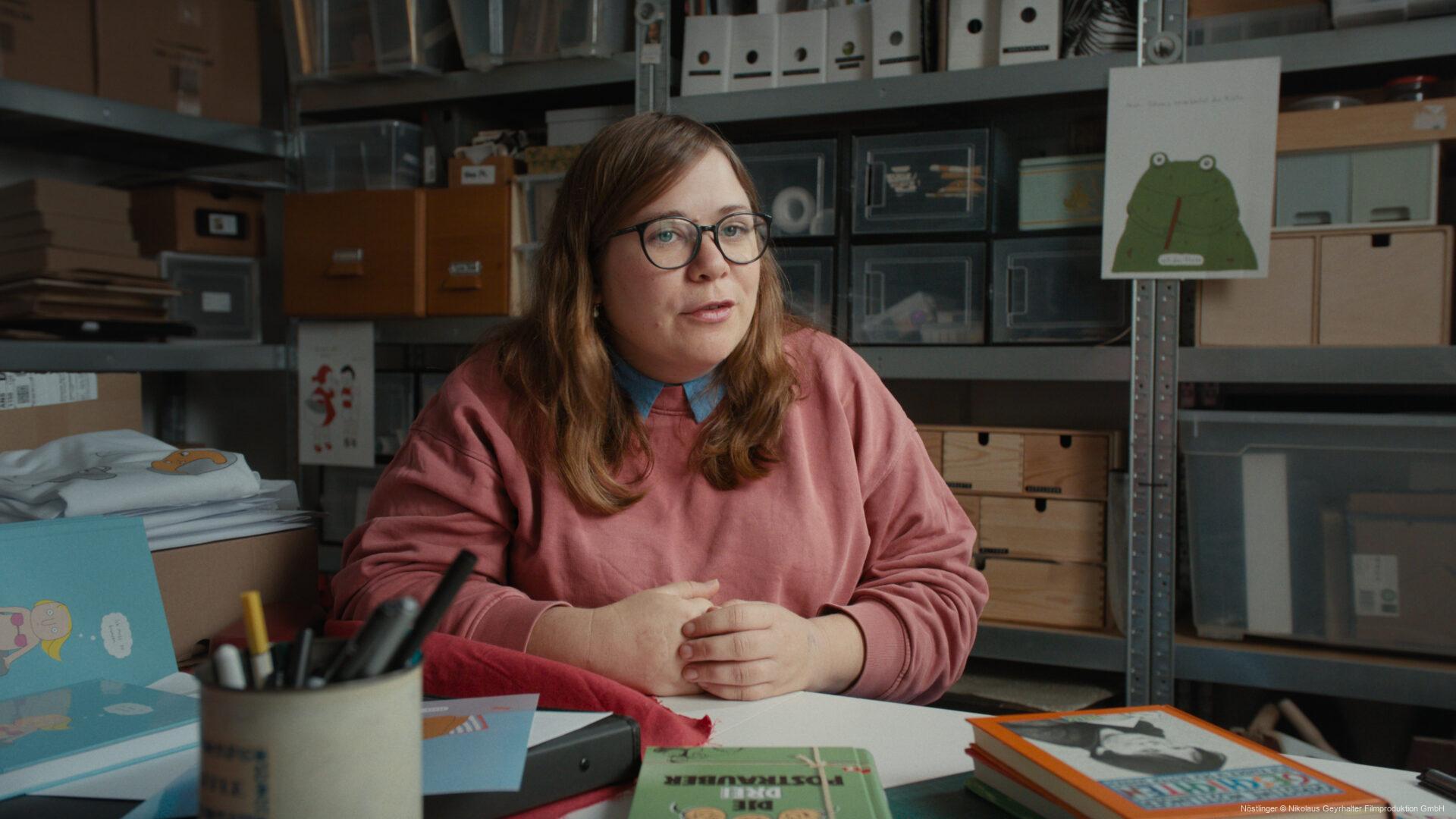 Frau mit Brille sitzt an einem Schreibtisch in einem Raum voller Regale, Kartons und Bürobedarf